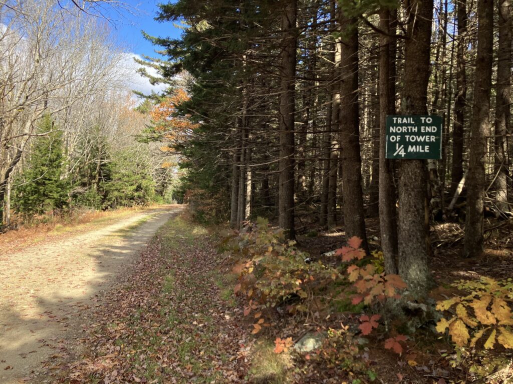 A dirt road leads through autumn woods. A sign attached to a tree to the right of the road reads "Trail to North end of tower 1/4 mile."