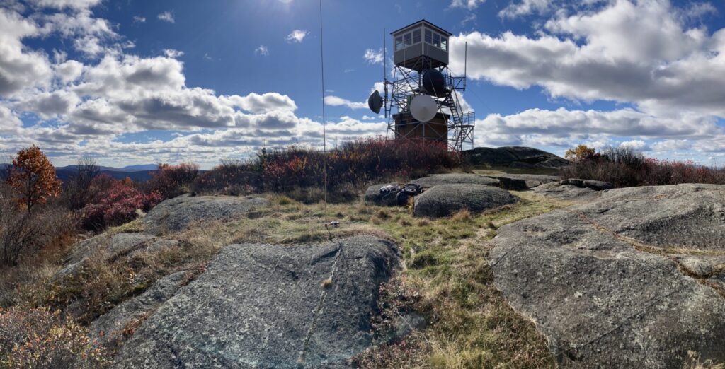 Panoramic photo of the top of Pitcher Mountain, showing low gray bedrock outcrops and low brush / scrub under a deep blue sky scattered with low, puffy clouds. A big mountain (Monadnock) looms in the distance. A short fire tower is at the summit with various microwave and other antennas clamped to it. In the middle ground of the photo, in a grassy spot between outcrops, is a ~18 ft tall vertical amateur radio antenna. It is connected by a cable snaking along the ground to a backpack and other bags resting on the edge of one of the rock outcrops.